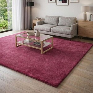 May include: A large, rectangular, deep magenta area rug with a plush, textured pile surface. The rug is positioned on a light-toned wooden floor, extending from the foreground toward a gray sofa, a glass coffee table, and a wooden side cabinet.