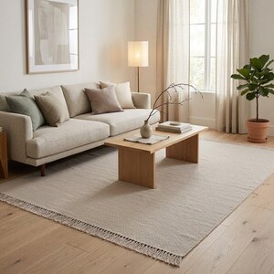 Könnte beinhalten: Ein beiges Sofa mit salbeigrünen und braunen Zierkissen, ein Couchtisch aus hellem Holz mit einer kleinen Vase und Büchern, eine Stehlampe, ein gerahmter abstrakter Druck und ein großer beiger Teppich mit Fransen auf hellem Holzboden.