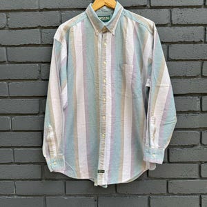 Può includere: Camicia a maniche lunghe a righe verticali nei colori pastello blu, viola, beige e bianco. La camicia ha un colletto button-down, una tasca sul petto e polsini con bottoni. La camicia è appesa a una gruccia di legno.