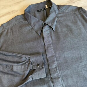 May include: A long-sleeved button-down shirt in a dark grey micro-patterned fabric. The shirt features a pointed collar, a front placket with buttons, and a label at the neckline that reads Versace Classic V2. The cuffs are secured with a single button.