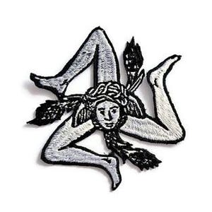 Puede incluir: Parche bordado con la Trinacria, un símbolo de Sicilia. El diseño representa una cabeza de mujer con tres piernas y espigas de trigo. El parche es principalmente blanco y negro, con costuras detalladas.