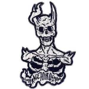May include: Embroidered patch featuring a skeletal demon with horns, a skull-like face, and the text "EMBRACE YOUR DEMONS" in a stylized font. The design is primarily black and white.