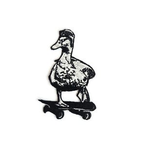 Könnte beinhalten: Schwarz-weißer, gestickter Aufnäher mit einer Ente auf einem Skateboard. Die Ente trägt eine Kappe. Das Skateboard ist ebenfalls schwarz. Das Design ist detailliert und strukturiert, ideal für Kleidung oder Accessoires.