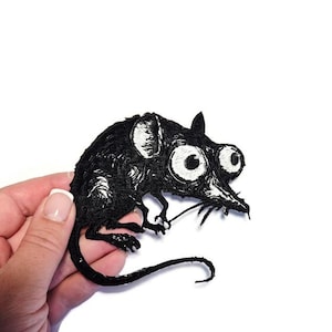 Puede incluir: Parche bordado negro de una rata de dibujos animados con ojos grandes y amplios y una larga cola. La rata tiene detalles blancos en las orejas y los ojos, y bigotes. El parche se sostiene sobre un fondo blanco.