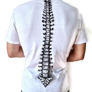 Puede incluir: Camiseta blanca con un diseño de columna vertebral en blanco y negro en la espalda. El diseño de la columna vertebral es detallado y recorre verticalmente el centro de la camiseta. La camiseta tiene mangas cortas y cuello redondo.