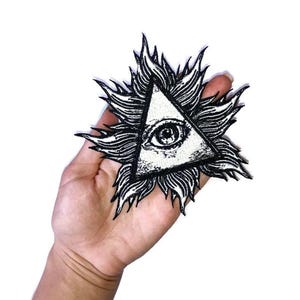 May include: Black and white embroidered patch featuring an eye within a triangle, surrounded by stylized rays. The patch is held in a hand against a white background. The design is a symbol of the all-seeing eye.
