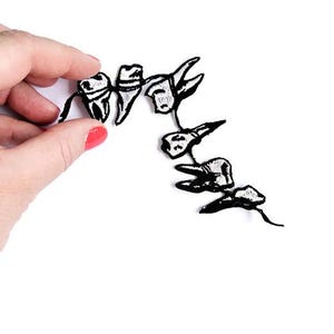 May include: Black and white embroidered teeth patches. The patches are in a line, with each tooth having a unique shape. The patches are held by a hand with red nail polish against a white background.