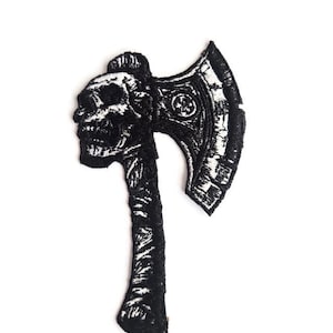 May include: Black and white embroidered patch in the shape of a skull-headed axe. The axe head features a skull design, and the handle is textured. The patch is likely for clothing or accessories.