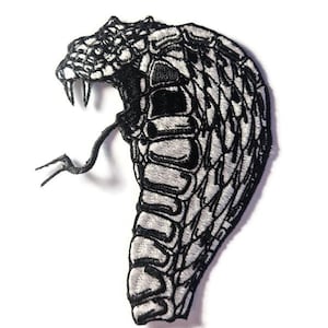 Puede incluir: Parche bordado que representa una cabeza de cobra con la boca abierta, revelando colmillos y una lengua bífida. El parche es principalmente blanco y negro, con costuras detalladas que crean una apariencia texturizada. La cabeza de la serpiente está angulada, dándole un aspecto dinámico.