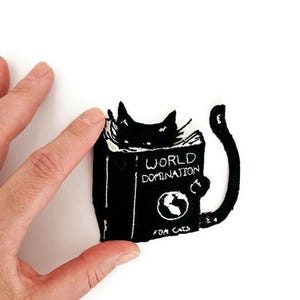 Könnte beinhalten: Schwarzer Katzenaufnäher mit dem Text "WORLD DOMINATION" und "FOR CATS" auf einem Buch. Die Katze schaut über das Buch. Der Aufnäher wird von einer Hand vor weißem Hintergrund gehalten.