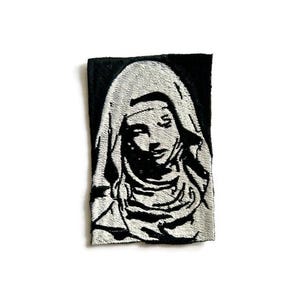 May include: Embroidered patch depicting a religious figure in black and white. The image shows a woman with a draped head covering, rendered in a detailed, stylized design. The patch has a black background.
