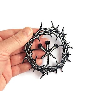 May include: A black and white embroidered patch featuring a crown of thorns encircling two crossed nails. The patch is held in a hand against a white background. The design is a religious symbol.