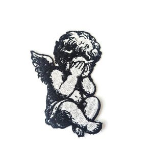 Può includere: Toppa ricamata in bianco e nero raffigurante un cherubino che si copre il viso. Il cherubino ha ali dettagliate e capelli ricci. La toppa è su sfondo bianco.