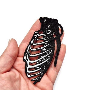 Pode incluir: Patch bordado a preto e branco em forma de granada com um design de caixa torácica de esqueleto. O patch é segurado em uma mão contra um fundo branco. O patch é um acessório único.