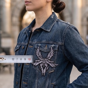 May include: A dark denim jacket with a large, black and white embroidered patch featuring a stylized winged design. The patch measures approximately 10 cm wide. The patch is on the left chest of the jacket.