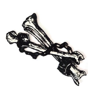 Puede incluir: Parche en blanco y negro con una representación estilizada de huesos cruzados. El parche tiene un aspecto texturizado, con los huesos representados en un estilo artístico detallado. El diseño está sobre un fondo blanco liso.