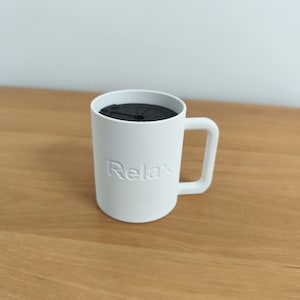 Puede incluir: Una taza blanca mate con un asa cuadrada y la palabra Relax grabada en relieve en la parte frontal. La taza cuenta con una tapa circular negra con un patrón radial que cubre la abertura superior, creando un diseño limpio y funcional para este recipiente.