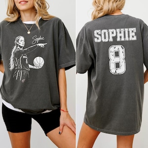 May include: A charcoal gray short-sleeve t-shirt. The front features a white graphic of a basketball player holding a ball with the name Sophie written in script. The back displays the name SOPHIE in white block letters above the number 8.