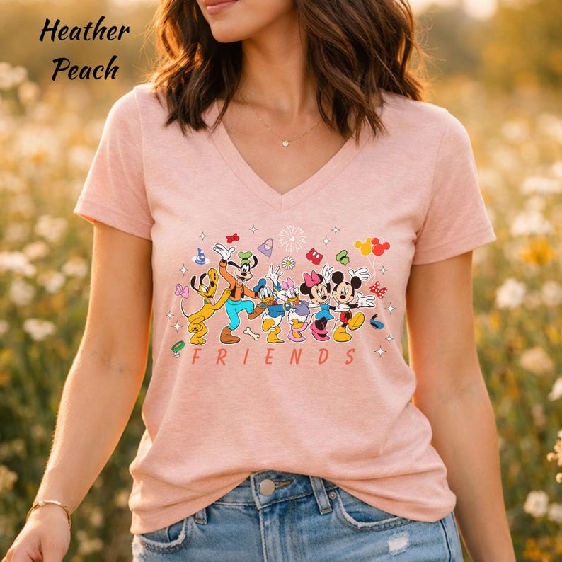 May include: A heather peach v-neck t-shirt featuring a graphic print of Disney characters including Pluto, Goofy, Donald Duck, Daisy Duck, Minnie Mouse, and Mickey Mouse. The word FRIENDS is printed in red below the characters with small icons.