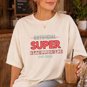 May include: An off-white short-sleeve t-shirt featuring the text ARTIFICIAL in light blue with a line through it, the word SUPER in large red letters, the word INTELLIGENCE in red outline, and the text EST.2026 in light blue at the bottom.