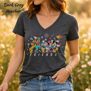May include: A dark grey heather V-neck t-shirt featuring a graphic print of Disney characters including Pluto, Goofy, Donald Duck, Daisy Duck, Minnie Mouse, and Mickey Mouse. The word FRIENDS is printed in white below the colorful character illustration.
