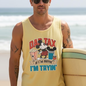 May include: Yellow tank top with a graphic featuring Mickey Mouse, popcorn, and a drink. The text reads "DAD TAX" above Mickey and "IF I'M BUYIN' I'M TRYIN'" below. The tank top is worn by a person holding a surfboard.