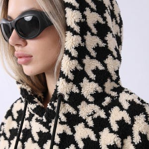 May include: A black and cream houndstooth patterned hoodie made of a textured, fuzzy fleece material. The garment features a hood with black drawstring cords and metal eyelets, providing a soft, thick appearance with a repeating geometric design.