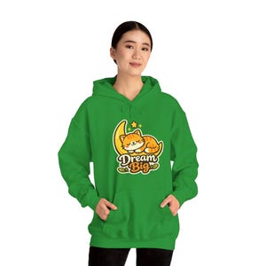 Peut inclure: Un sweat à capuche vert vif avec un chat de dessin animé dormant sur un croissant de lune et une étoile. Le texte "Dream Big" est en dessous du chat. Le sweat à capuche a une poche avant et une capuche à cordon.