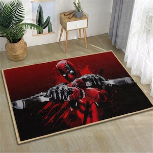 May include: Rectangular area rug featuring a graphic of Deadpool in a red and black color scheme. The rug has a tan border and depicts Deadpool holding two guns against a red background with black accents. The rug is suitable for a living room or bedroom.