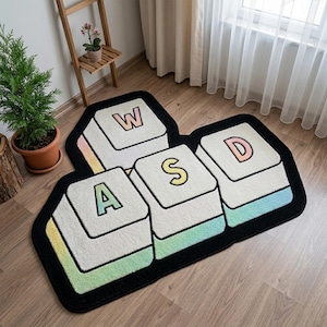 May include: A floor rug shaped like four computer keyboard keys arranged in a WASD configuration. The keys are white with pastel-colored letters and edges, outlined in a thick black border. The rug features the letters W, A, S, and D.