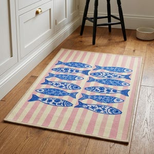 May include: A rectangular rug with a pink and cream striped background. The rug features a pattern of blue fish with floral and geometric designs. The rug is placed on a wooden floor, suggesting a kitchen or dining area.