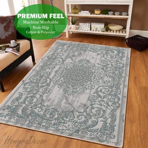 May include: A rectangular area rug with a gray and teal floral pattern. The rug has a central medallion design and a border with scrolling leaves. The rug is made of cotton and polyester and is machine washable. The text "PREMIUM FEEL" is visible.