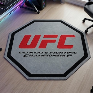 Peut inclure: Tapis octogonal gris avec le logo UFC en rouge et l'inscription "ULTIMATE FIGHTING CHAMPIONSHIP" en noir. Le tapis a une bordure noire et est posé sur un sol en bois clair.