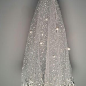 Puede incluir: Un velo blanco y transparente con lentejuelas dispersas y perlas grandes y redondas. El velo se sujeta con un hilo de perlas. El borde inferior está adornado con más perlas, creando un aspecto delicado y elegante. Probablemente para una boda u ocasión especial.