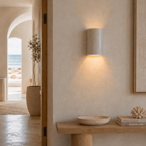 May include: A cylindrical wall sconce with a smooth, light grey finish mounted on a beige wall, emitting a warm, downward-facing light. The fixture features a simple, curved design that directs illumination towards the surface below it.