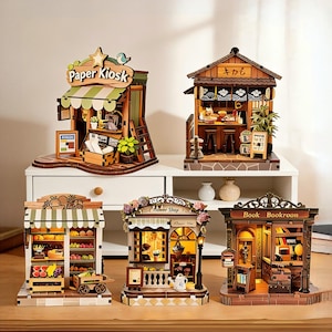 May include: Five miniature wooden shop models including a Paper Kiosk, a Japanese-style shop with text, a fruit stand, a flower shop, and a book room. Each structure features detailed interiors, small furniture, and integrated warm lighting.