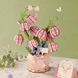 Puede incluir: Un kit de manualidades decorativo de madera con flores de lirio de los valles rosas y tallos verdes en una maceta rosa claro adornada con un lazo de satén. El arreglo incluye pequeñas flores azules y un letrero que dice Lily of the Valley.