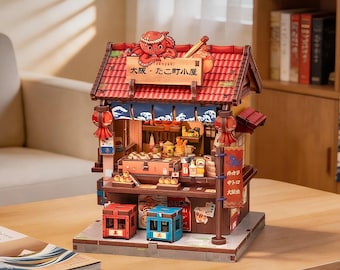 Puede incluir: Una maqueta de madera en miniatura de una tienda de takoyaki con techo de tejas rojas, una figura de pulpo y texto japonés en un letrero. El puesto incluye un mostrador con comida, dos taburetes pequeños en rojo y azul y farolillos.
