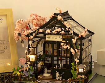 May include: A miniature model of a black-framed greenhouse with a glass roof, adorned with pink cherry blossom branches. The structure features a sign with the Japanese text 桜の花房, interior shelves, two cat figurines, and exterior lanterns.