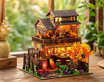 Puede incluir: Una maqueta de madera en miniatura de un restaurante japonés de dos plantas con tejas de color marrón oscuro, farolillos de papel iluminados, un balcón con barandilla de madera y un pequeño árbol con hojas de otoño naranjas. La estructura incluye asientos interiores detallados, letreros con caracteres japoneses y una pequeña zona de jardín con plantas en macetas y una linterna de piedra.