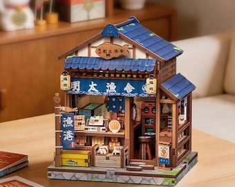 May include: A miniature wooden model of a Japanese fish market shop featuring a blue tiled roof, a blue fabric awning with white Japanese text, a wooden fish sign, and detailed interior displays including stools, crates, and small food items.