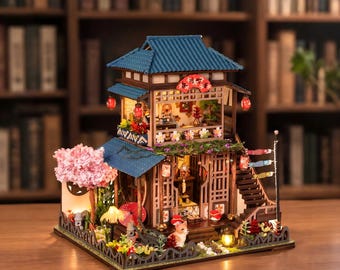 Puede incluir: Una maqueta de edificio de madera de dos plantas con tejado de tejas azules, un cerezo en flor rosa y farolillos iluminados. La estructura incluye una escalera, una pequeña figura de perro y elementos decorativos como banderas de carpas.