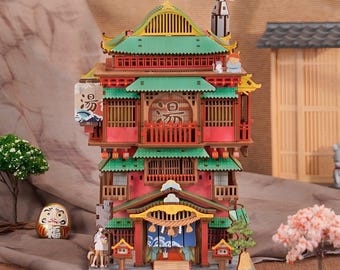 May include: A multi-story wooden model of a traditional Japanese bathhouse featuring red walls, green tiled roofs, and intricate architectural details. The structure includes a chimney with smoke, a hanging lantern with Japanese text, and a small figurine.