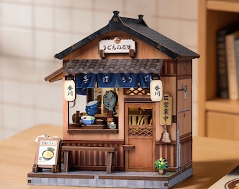 Puede incluir: Una maqueta de madera en miniatura de una tienda de fideos udon japonesa con un tejado de tejas oscuras, una cortina noren azul con texto blanco, dos farolillos iluminados, una puerta de madera, un banco pequeño y un menú con un bol de fideos.
