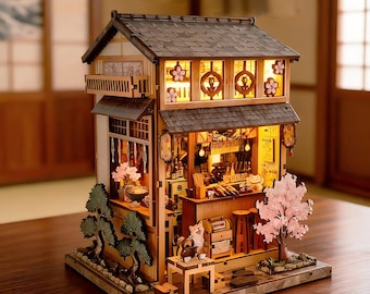 May include: A miniature wooden model of a two-story Japanese-style shop with a dark tiled roof, illuminated interior, and exterior details including a pink cherry blossom tree, two small pine trees, and a small table with a dog figurine in front.