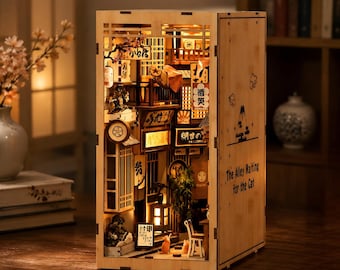 May include: A wooden book nook diorama featuring an illuminated miniature Japanese alleyway scene. The exterior side panel is engraved with the text The Alley Waiting for the Cat and a mountain graphic. The interior displays lanterns, signs, and cats.
