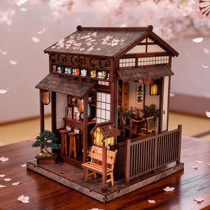 Könnte beinhalten: Ein Miniatur-Holzmodell eines japanischen Teehauses mit einem dunkelbraunen Ziegeldach, das mit rosa Blütenblättern bestreut ist, einem kleinen Bonsai-Baum, einer Holzbank und Innendetails, darunter eine Wandrolle mit dem japanischen Text 生の茶.
