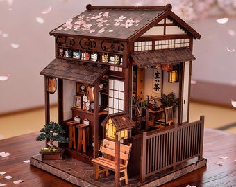 May include: A miniature wooden model of a Japanese tea house featuring a dark brown tiled roof with scattered pink petals, a small bonsai tree, a wooden bench, and interior details including a wall scroll with the Japanese text 生の茶.