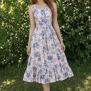Puede incluir: Un vestido de largo midi con una base de color crema y un estampado floral azul. La prenda tiene un escote cuadrado, tirantes anchos, un corpiño ajustado, una cintura fruncida y una falda terminada con un dobladillo de volantes.