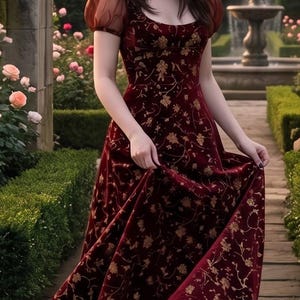 Puede incluir: Un vestido de terciopelo burdeos con bordados florales dorados. El vestido tiene mangas cortas y transparentes y un escote corazón. La mujer está en un jardín con una fuente y rosales.